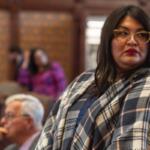 Woman with dark hair and glasses wearing a blue plaid shawl, looking to the right at a meeting.