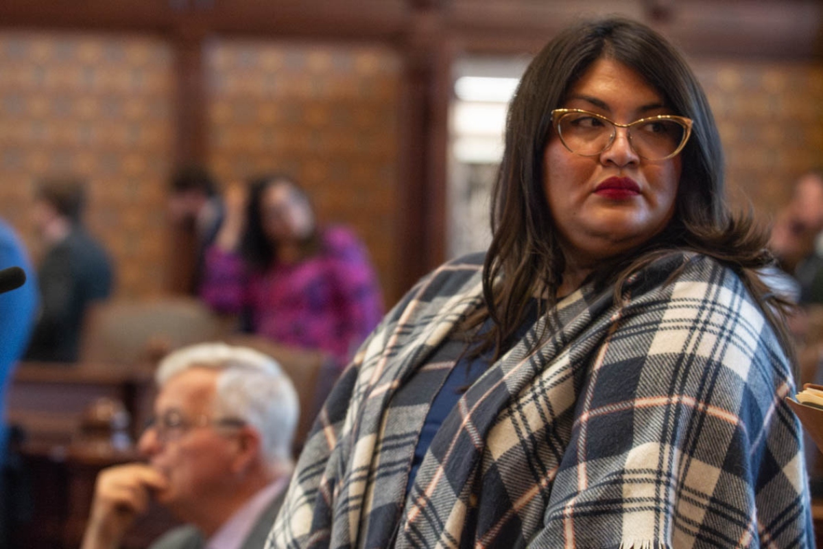 Woman with dark hair and glasses wearing a blue plaid shawl, looking to the right at a meeting.