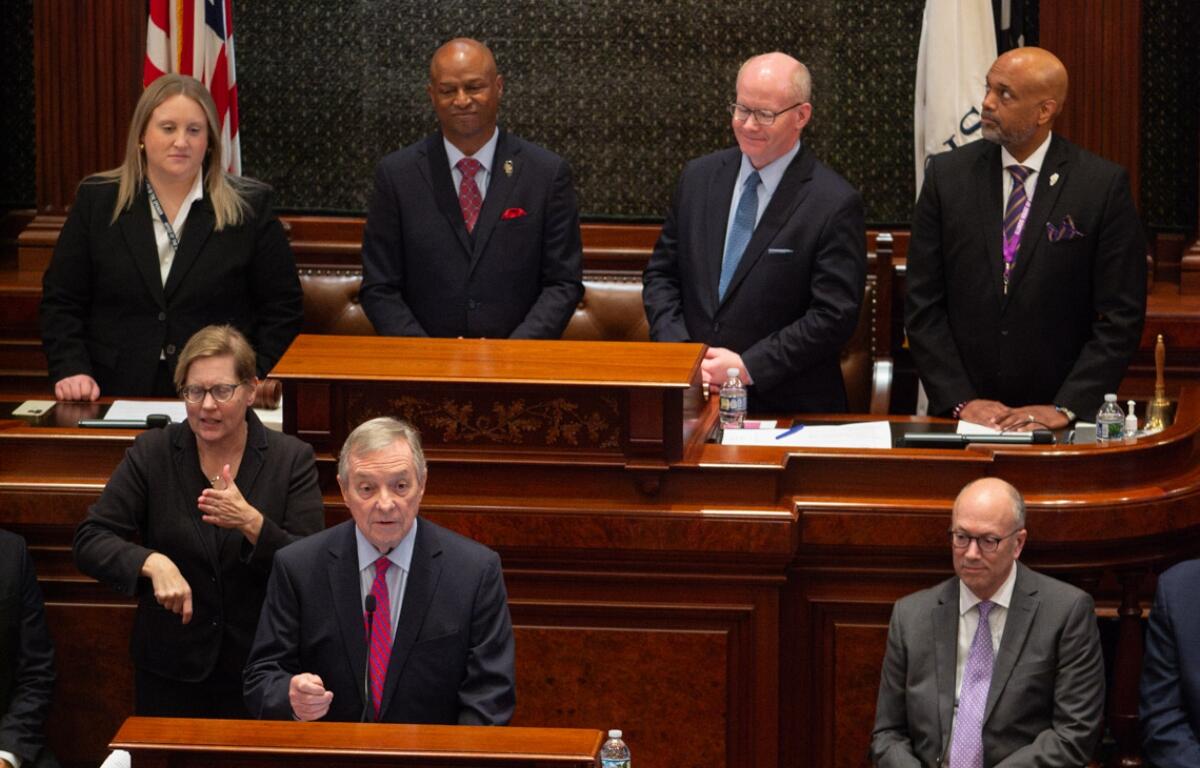 A man in a dark suit speaks at a wooden podium in a formal government chamber, with four officials standing behind him and a sign-language interpreter nearby.
