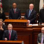 A man in a dark suit speaks at a wooden podium in a formal government chamber, with four officials standing behind him and a sign-language interpreter nearby.