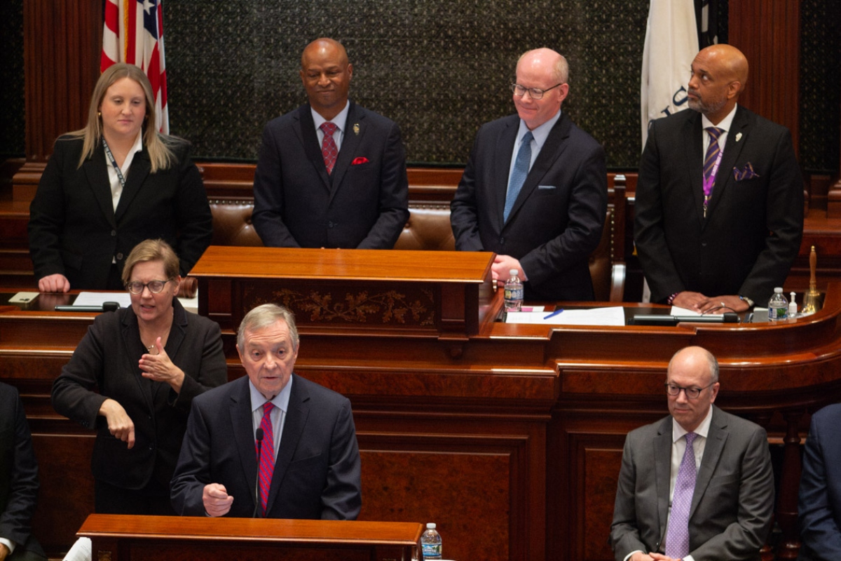 A man in a dark suit speaks at a wooden podium in a formal government chamber, with four officials standing behind him and a sign-language interpreter nearby.