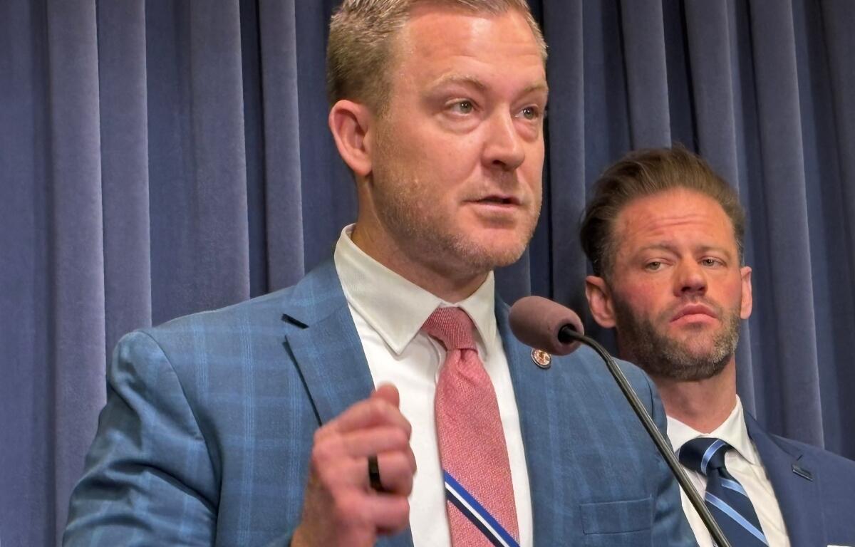 Speaker in a blue suit and pink tie at a podium with a microphone; a second man in a suit stands behind him on a stage.