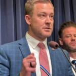 Speaker in a blue suit and pink tie at a podium with a microphone; a second man in a suit stands behind him on a stage.