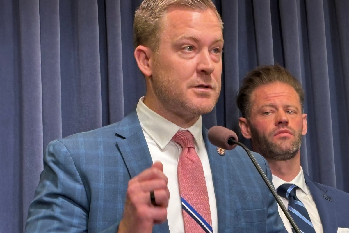 Speaker in a blue suit and pink tie at a podium with a microphone; a second man in a suit stands behind him on a stage.