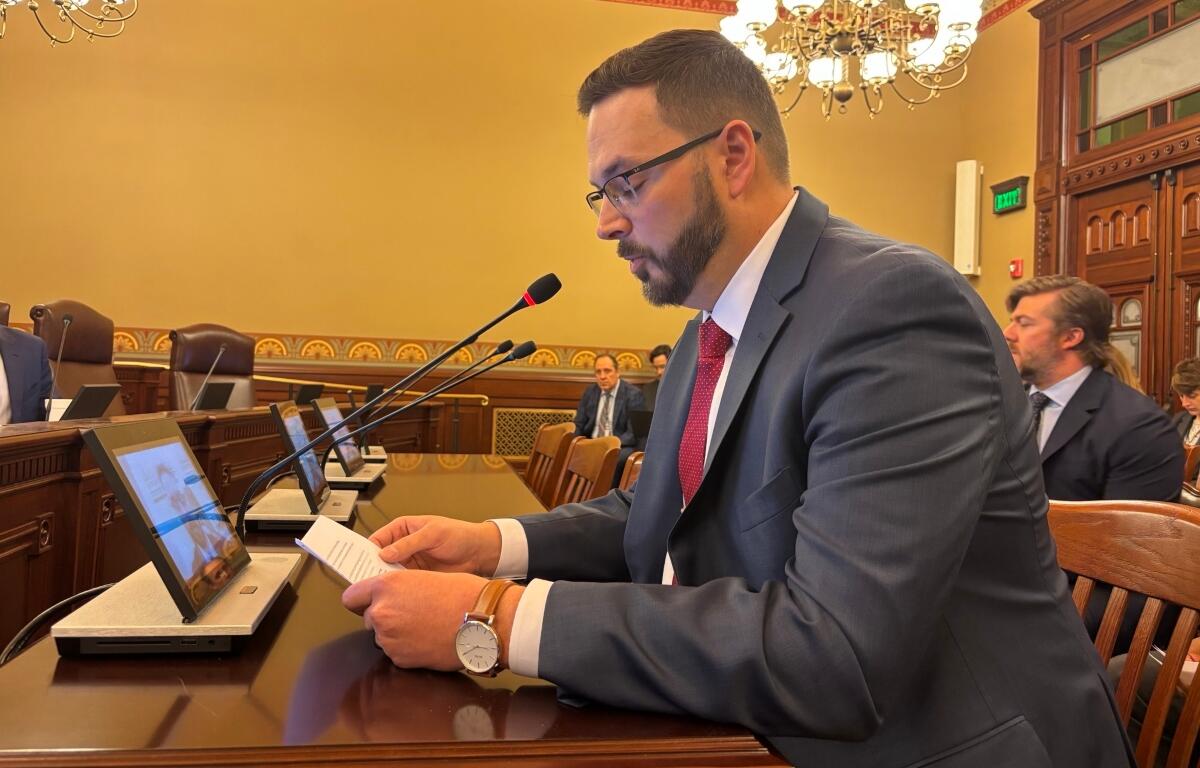 Man in a suit and red tie sits at a conference desk, reading notes into a microphone during a formal meeting.