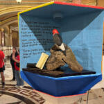 Blue exhibiti​on case with a carved log and a red-headed woodpecker perched on it, an open book, and handwritten text on a blue backdrop; students in red shirts nearby.