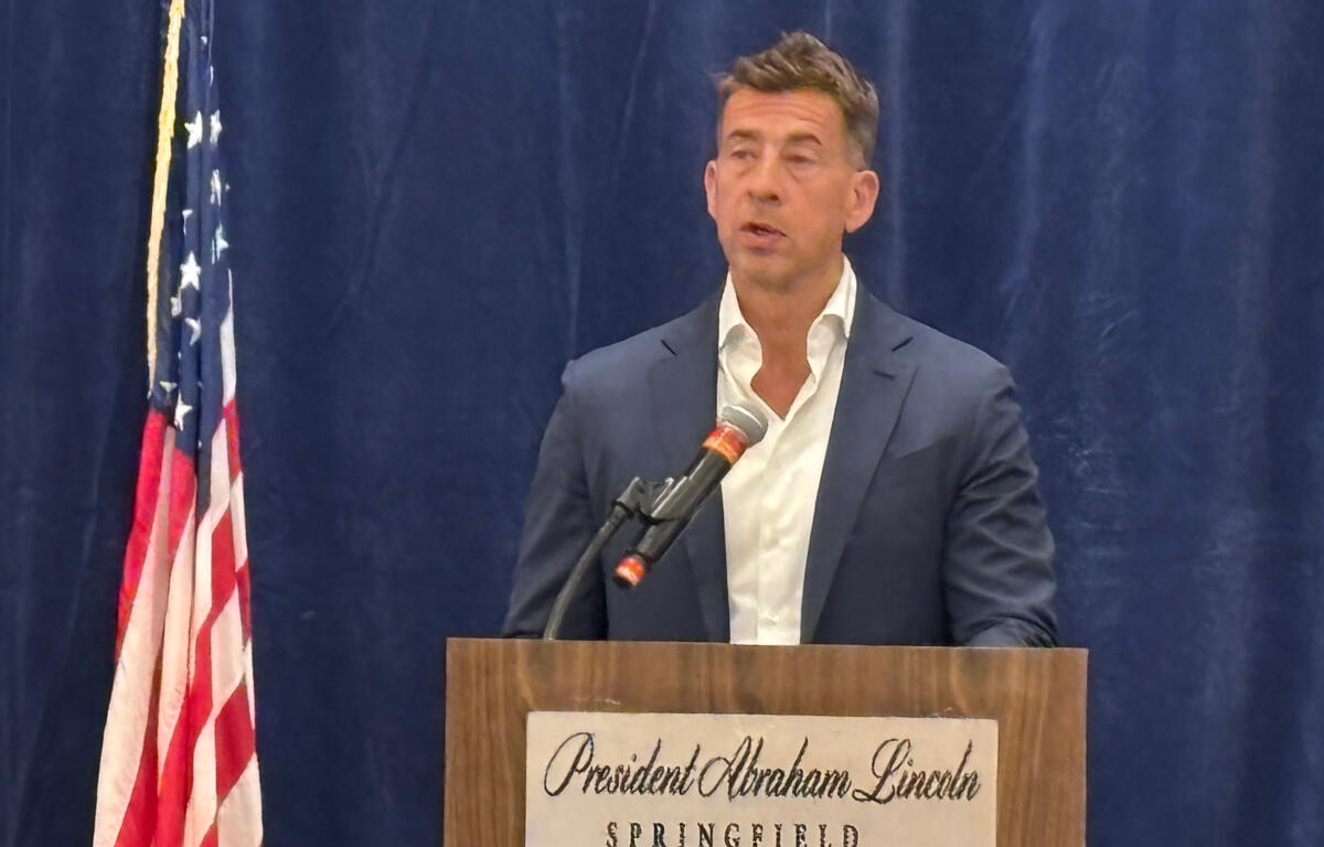 Man in a navy suit delivers a speech at a wooden podium, with an American flag to the left and a blue curtain backdrop.
