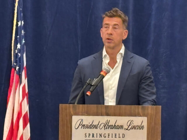 Man in a navy suit delivers a speech at a wooden podium, with an American flag to the left and a blue curtain backdrop.