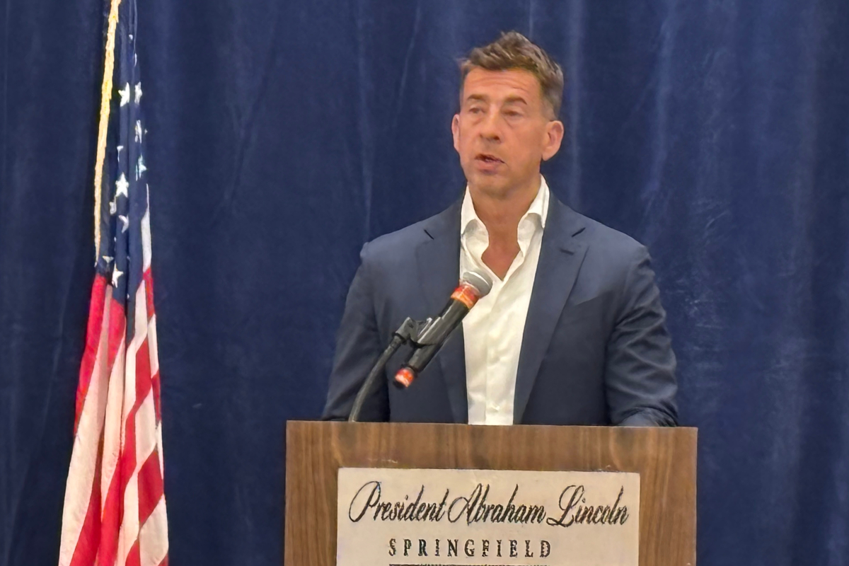 Man in a navy suit delivers a speech at a wooden podium, with an American flag to the left and a blue curtain backdrop.