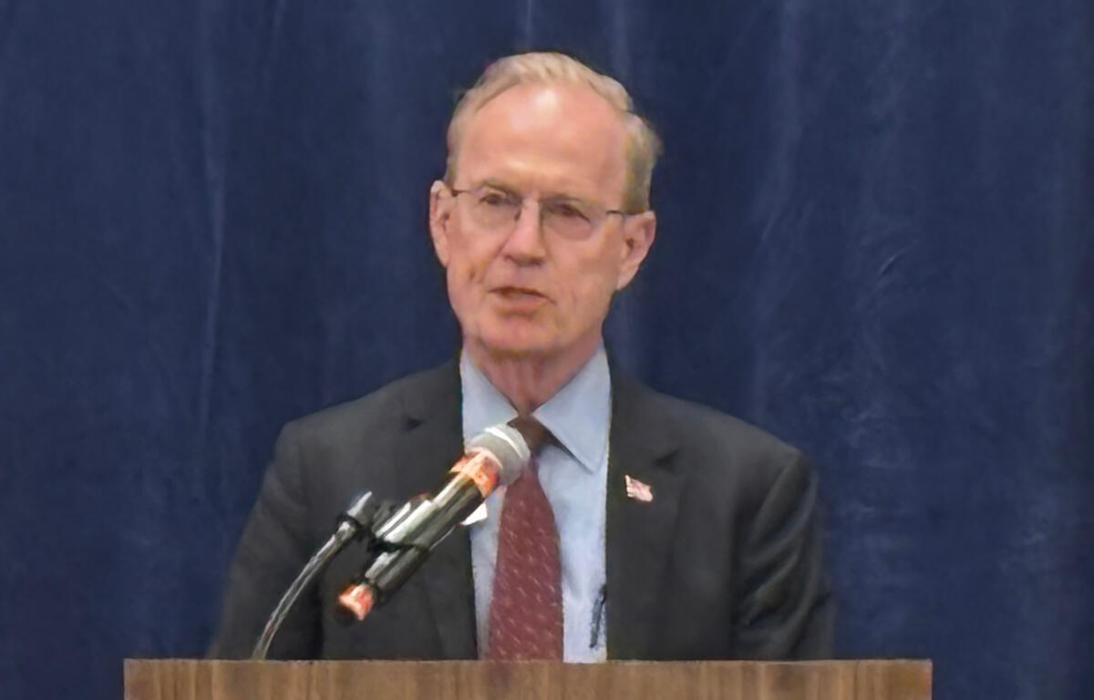 Man in a dark suit and red tie speaking at a wooden podium with a microphone, stands before a blue curtain.