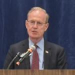 Man in a dark suit and red tie speaking at a wooden podium with a microphone, stands before a blue curtain.