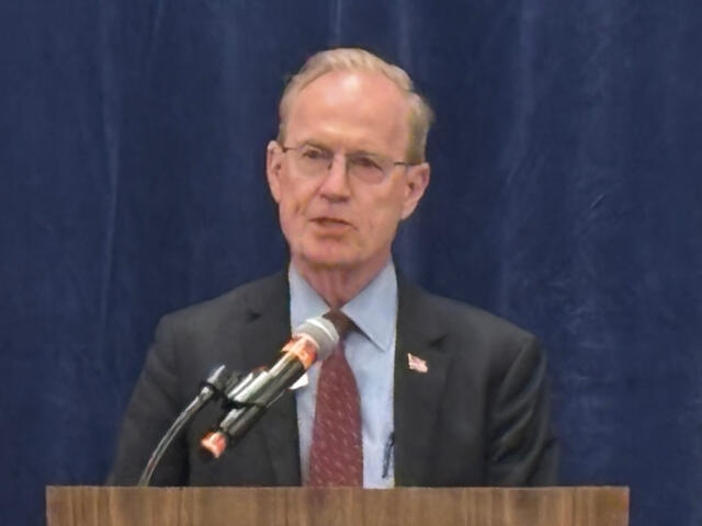 Man in a dark suit and red tie speaking at a wooden podium with a microphone, stands before a blue curtain.
