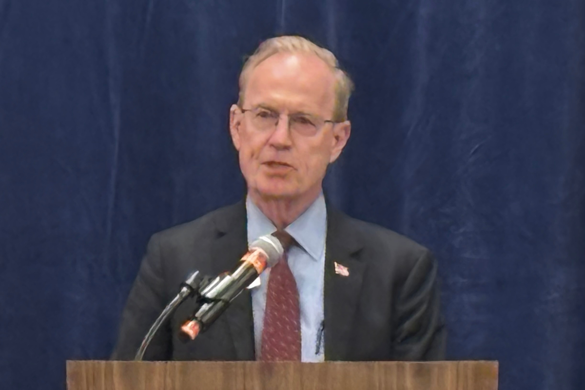 Man in a dark suit and red tie speaking at a wooden podium with a microphone, stands before a blue curtain.