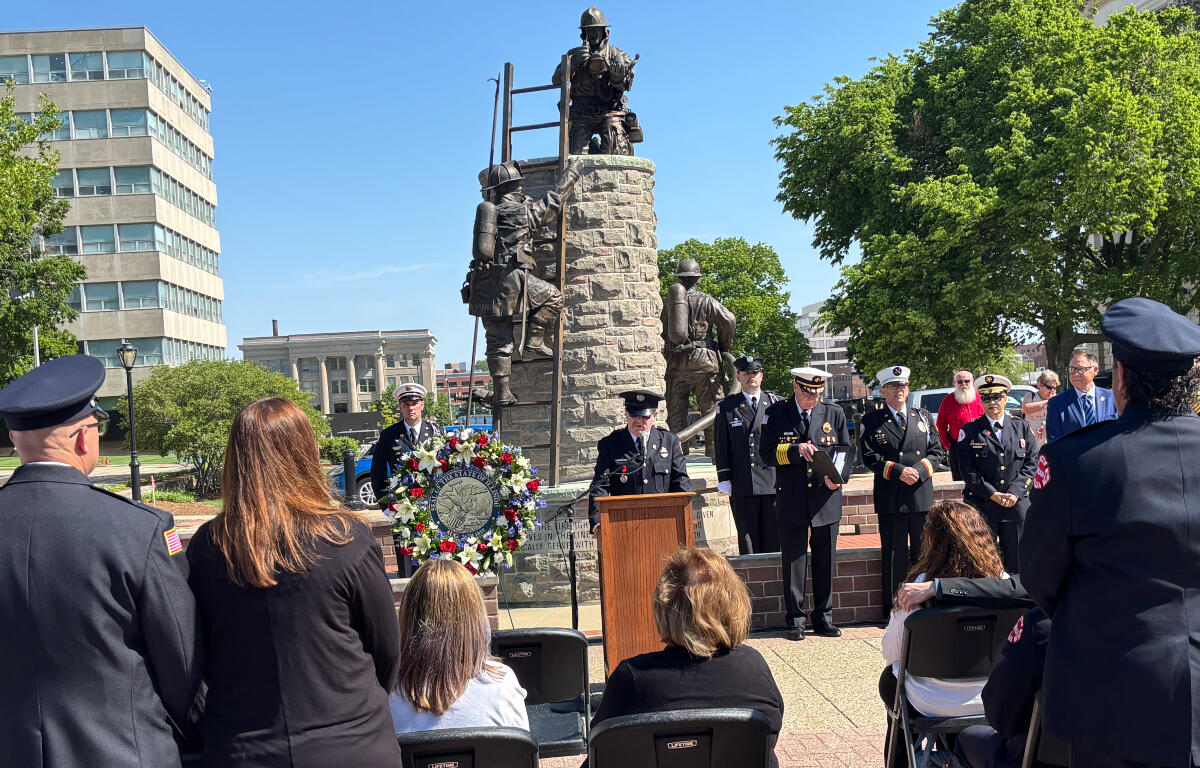 Ceremony at a memorial statue with uniformed attendees, a wreath, and a speaker at a podium.