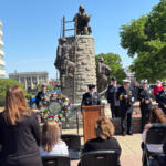 Ceremony at a memorial statue with uniformed attendees, a wreath, and a speaker at a podium.