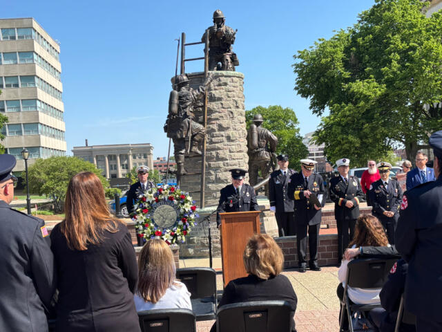 Ceremony at a memorial statue with uniformed attendees, a wreath, and a speaker at a podium.