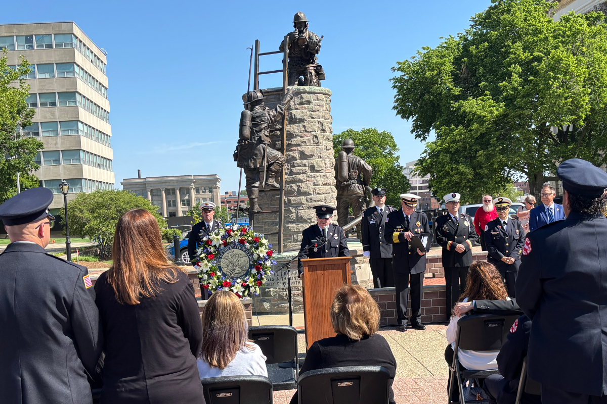 Ceremony at a memorial statue with uniformed attendees, a wreath, and a speaker at a podium.