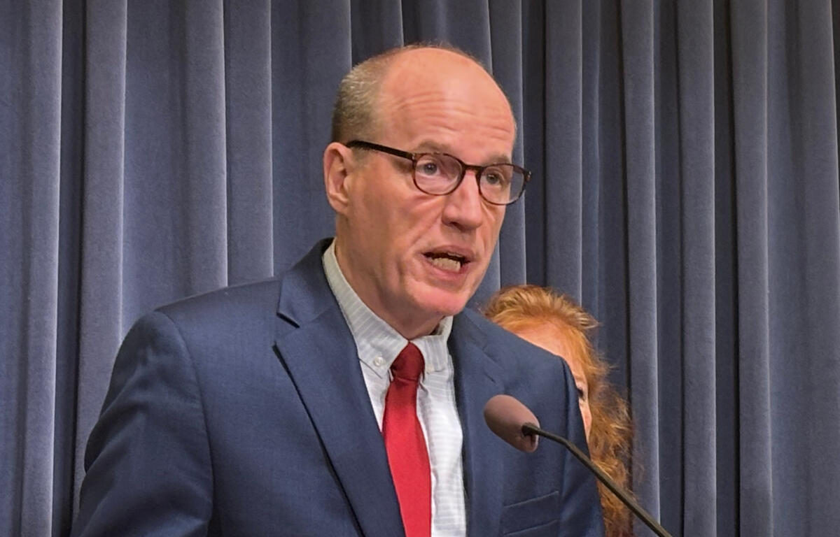 Man in a navy suit and red tie speaks into a microphone in front of blue curtains.