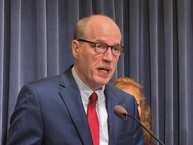 Man in a navy suit and red tie speaks into a microphone in front of blue curtains.