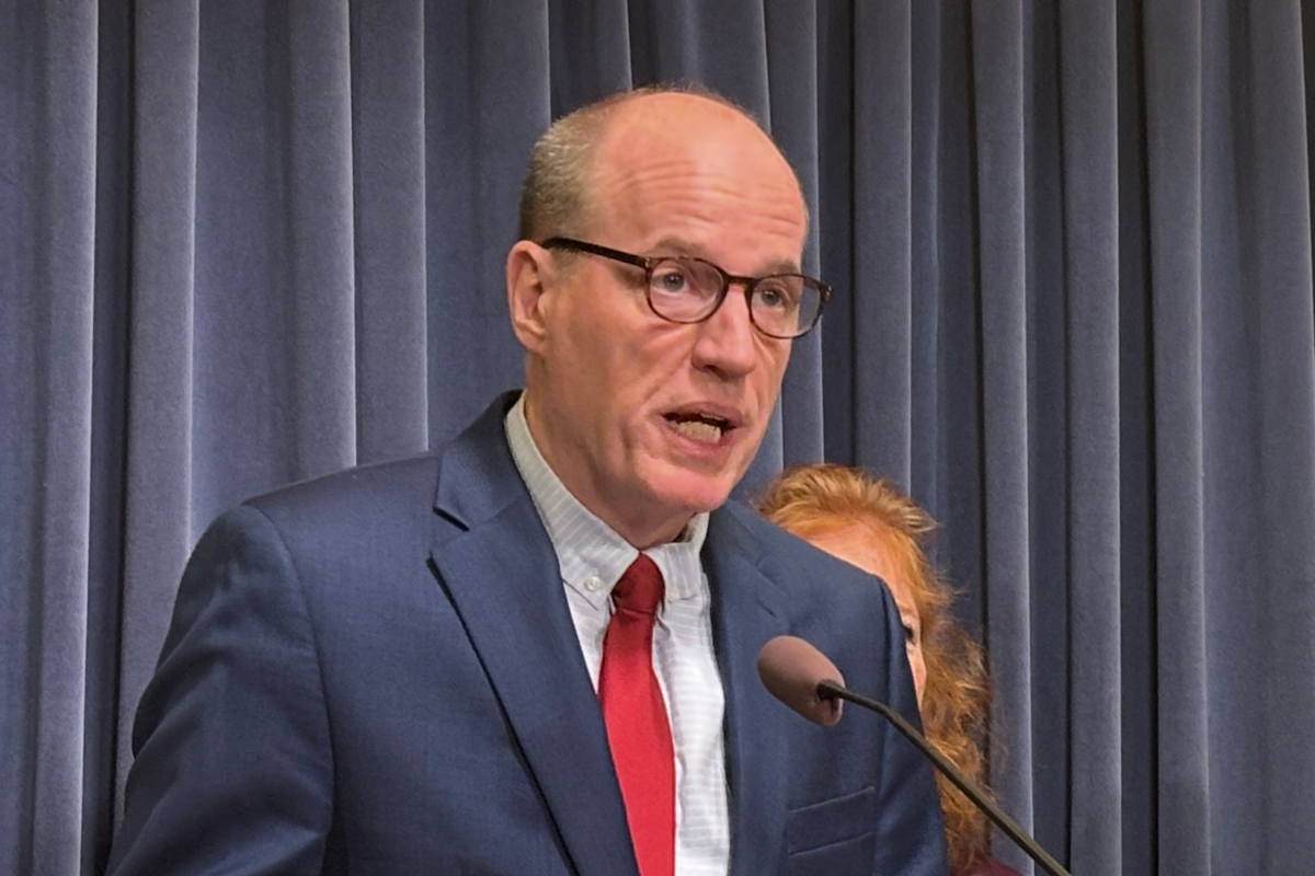 Man in a navy suit and red tie speaks into a microphone in front of blue curtains.
