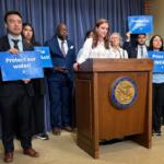 Diverse group at a press conference behind a podium, holding signs that urge Protect our water and Pass the POWER Act.