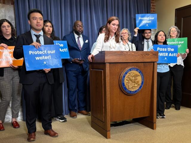 Diverse group at a press conference behind a podium, holding signs that urge Protect our water and Pass the POWER Act.