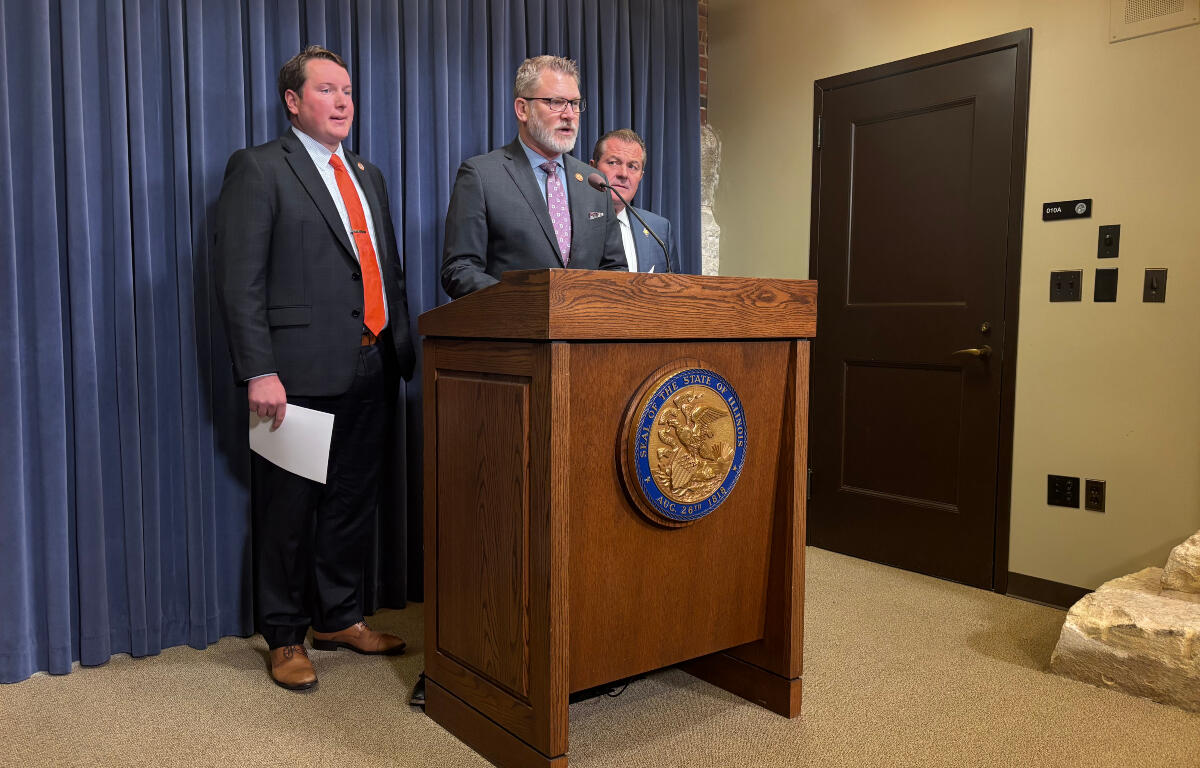 Man in a suit speaks at a wooden podium with a state seal, flanked by two other men in suits against a blue curtain backdrop.