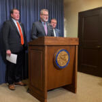 Man in a suit speaks at a wooden podium with a state seal, flanked by two other men in suits against a blue curtain backdrop.