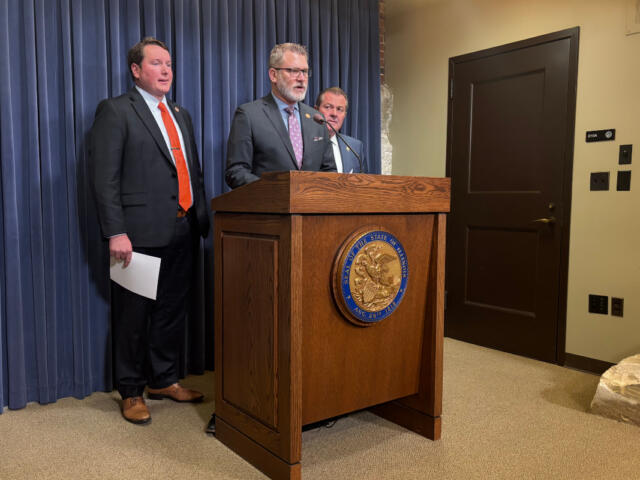 Man in a suit speaks at a wooden podium with a state seal, flanked by two other men in suits against a blue curtain backdrop.