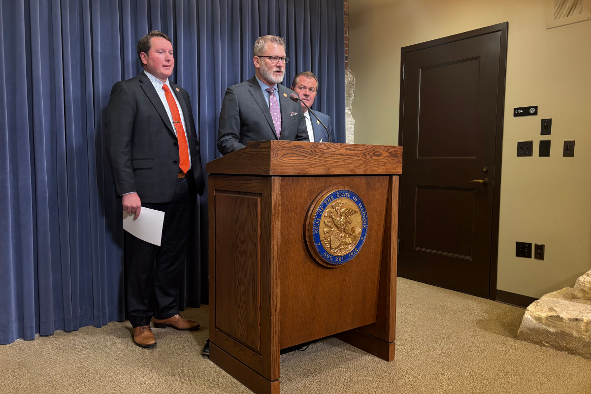 Man in a suit speaks at a wooden podium with a state seal, flanked by two other men in suits against a blue curtain backdrop.