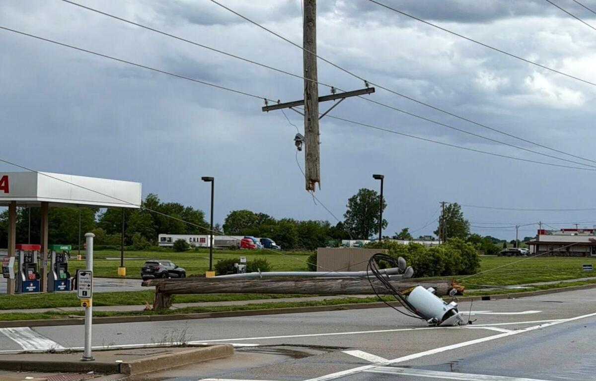 Downed wooden utility pole with tangled power lines across a street near a gas station, creating a hazard route barrier.
