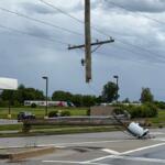 Downed wooden utility pole with tangled power lines across a street near a gas station, creating a hazard route barrier.