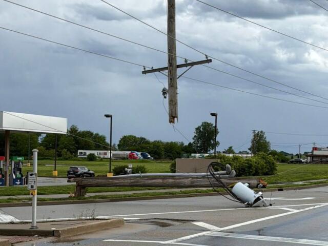 Downed wooden utility pole with tangled power lines across a street near a gas station, creating a hazard route barrier.