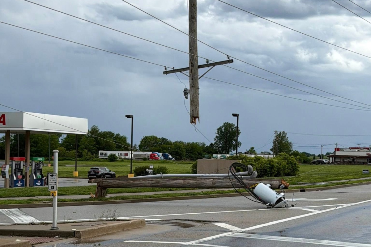 Downed wooden utility pole with tangled power lines across a street near a gas station, creating a hazard route barrier.