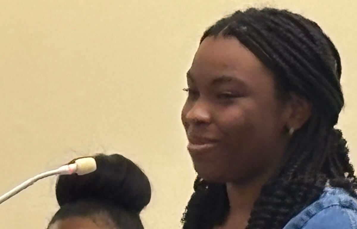 Woman with braided hair speaking into a microphone, wearing a blue top, in a neutral setting.