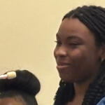 Woman with braided hair speaking into a microphone, wearing a blue top, in a neutral setting.