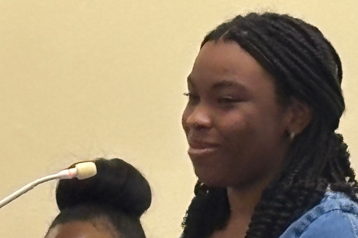 Woman with braided hair speaking into a microphone, wearing a blue top, in a neutral setting.