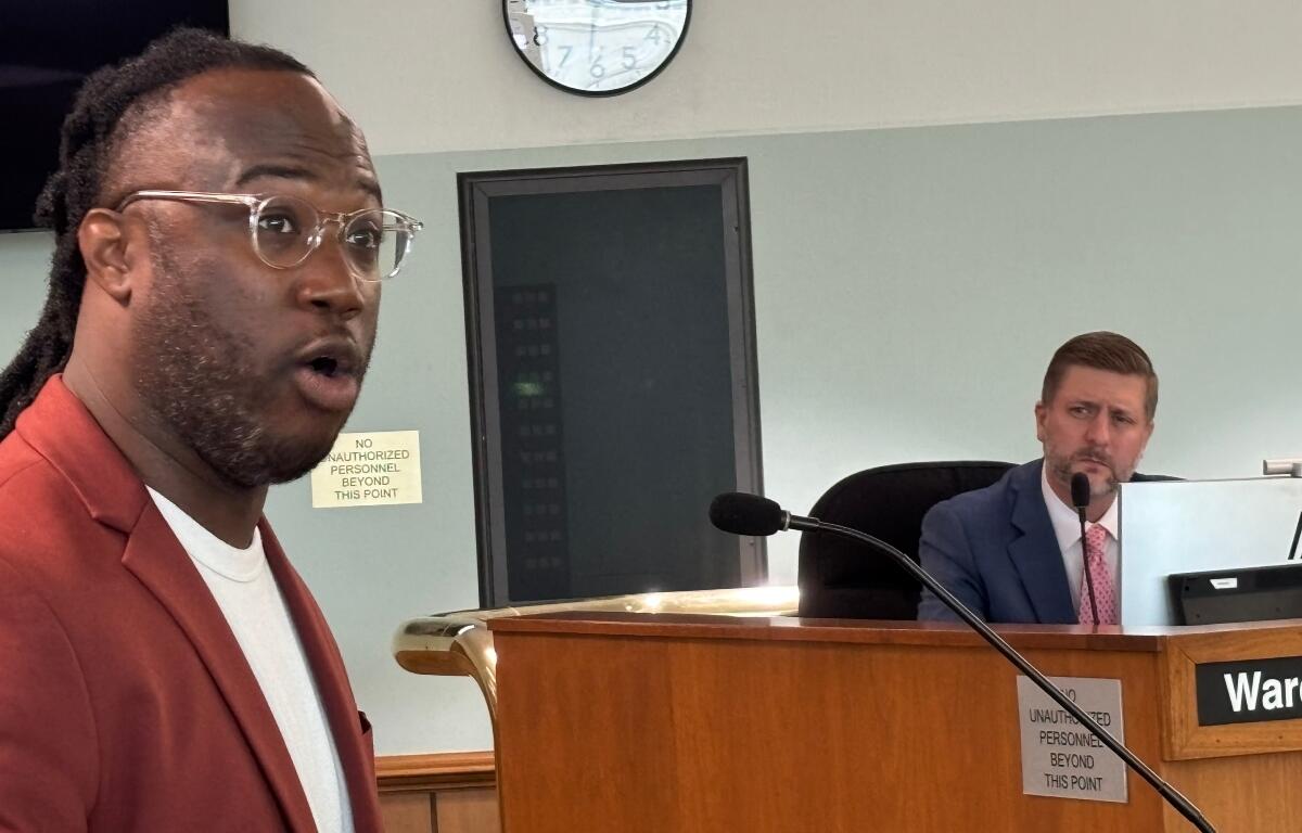 Man in a red blazer speaks toward the camera in a formal setting, a man in a suit seated at a desk behind him in a council/boardroom.