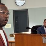 Man in a red blazer speaks toward the camera in a formal setting, a man in a suit seated at a desk behind him in a council/boardroom.