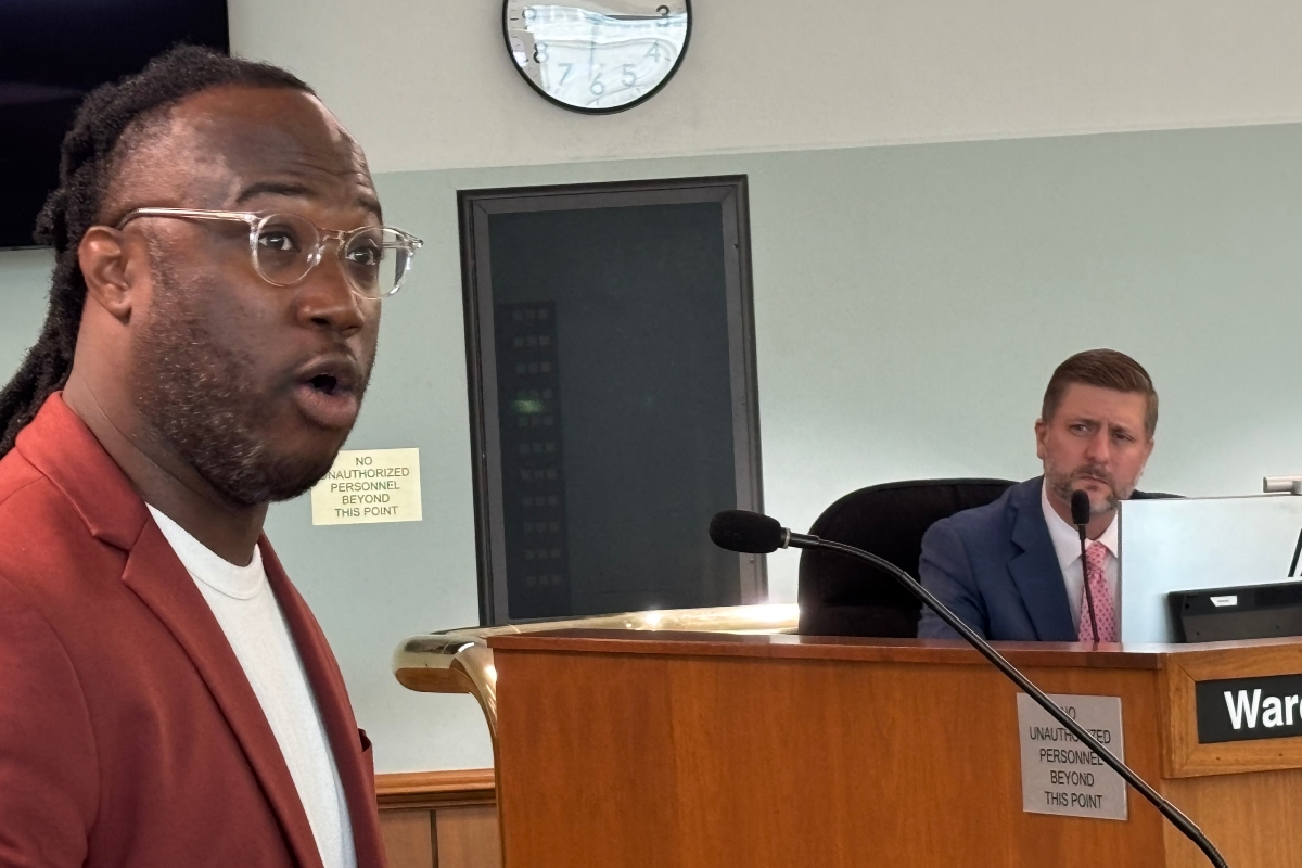 Man in a red blazer speaks toward the camera in a formal setting, a man in a suit seated at a desk behind him in a council/boardroom.