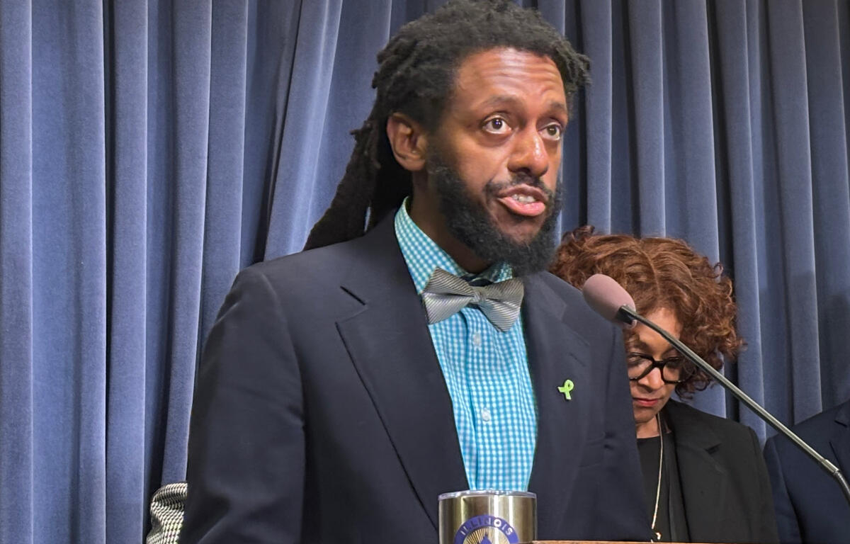 Speaker at a podium delivering a speech; wearing a navy blazer, teal check shirt, gray bow tie, and a green awareness ribbon, with blue curtains in the background.