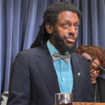 Speaker at a podium delivering a speech; wearing a navy blazer, teal check shirt, gray bow tie, and a green awareness ribbon, with blue curtains in the background.