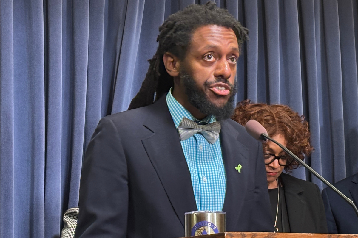 Speaker at a podium delivering a speech; wearing a navy blazer, teal check shirt, gray bow tie, and a green awareness ribbon, with blue curtains in the background.
