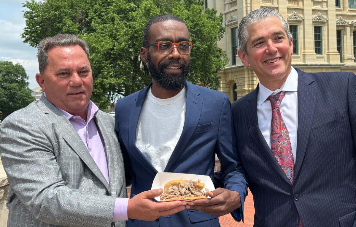 Three men in suits posing outdoors, holding a plate of sandwiches.
