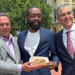Three men in suits posing outdoors, holding a plate of sandwiches.