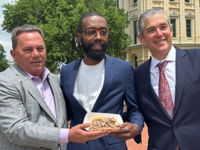 Three men in suits posing outdoors, holding a plate of sandwiches.