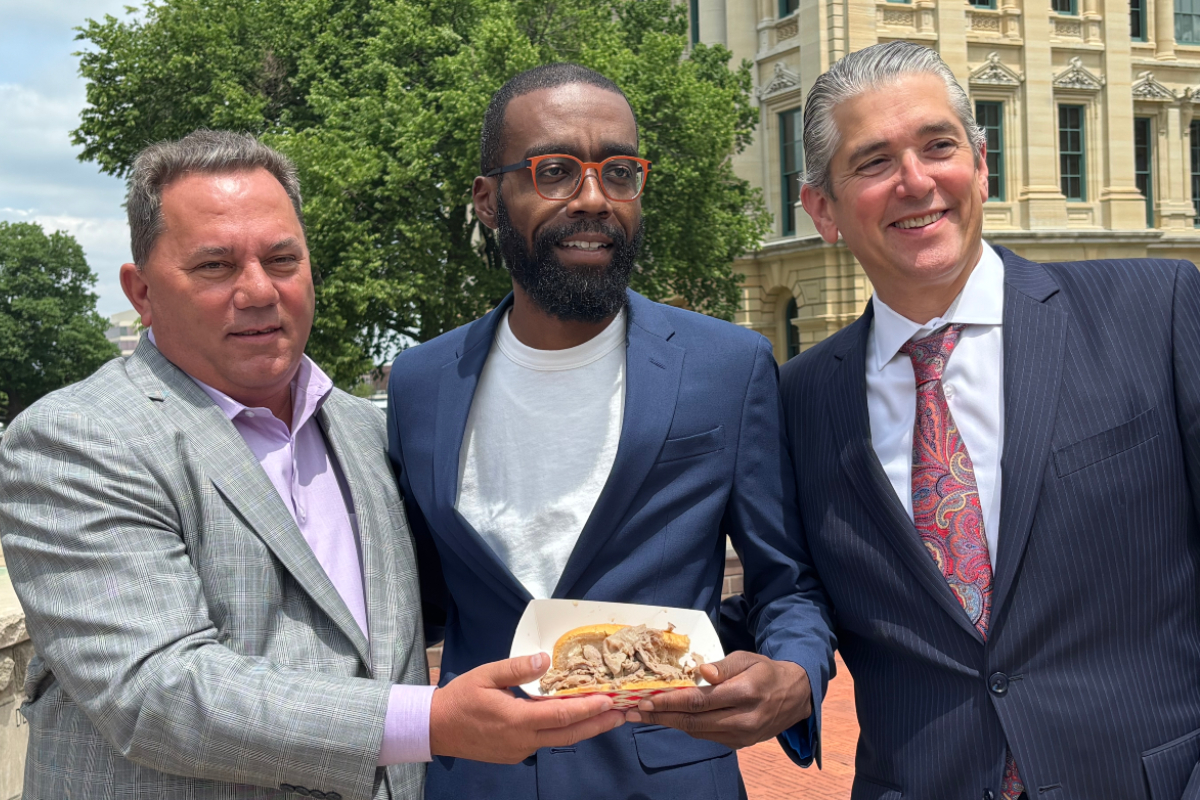 Three men in suits posing outdoors, holding a plate of sandwiches.