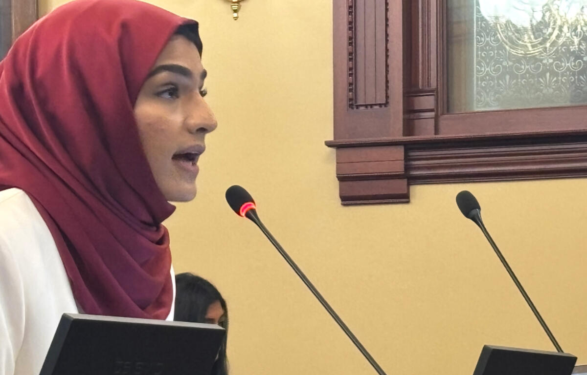 Profile of a woman in a maroon hijab speaking into a microphone at a lectern in a formal room, holding a folder.