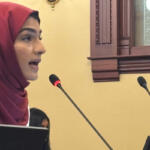 Profile of a woman in a maroon hijab speaking into a microphone at a lectern in a formal room, holding a folder.