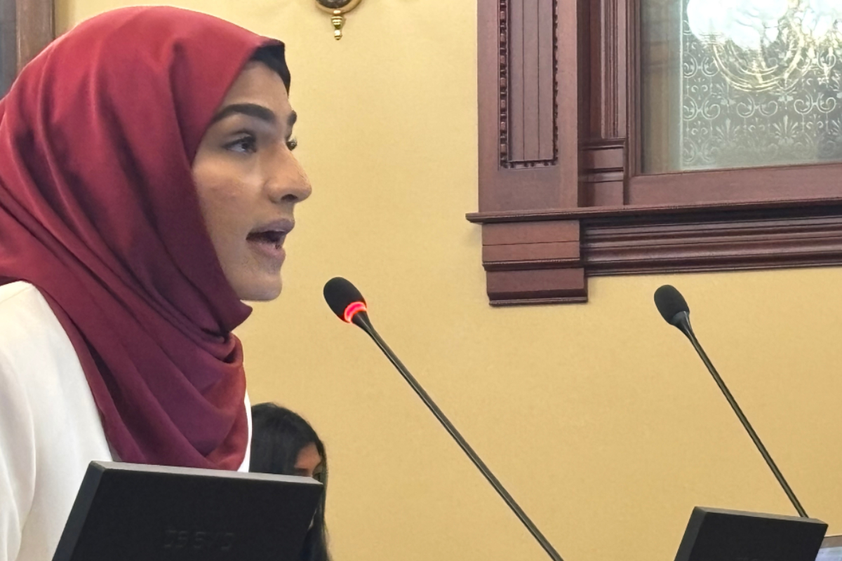 Profile of a woman in a maroon hijab speaking into a microphone at a lectern in a formal room, holding a folder.